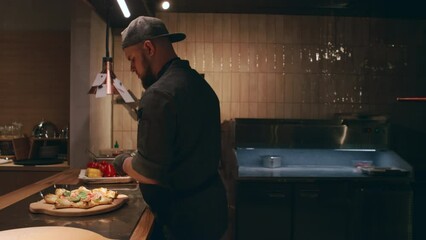 Medium side shot of young Caucasian male chef garnishing freshly baked pizza with basil leaves, then grabbing bottle from counter, shaking it and adding drops of sauce on top