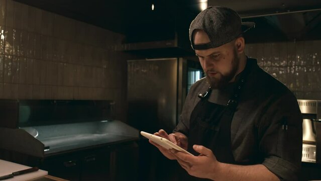 Medium tilting shot of Caucasian male chef in uniform, cap and apron walking up to counter at start of shift in restaurant kitchen, picking up tablet computer and checking for new orders