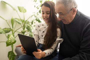 Cute little girl and her handsome grandpa are smiling while sitting on couch at home. Girl is using a tablet.