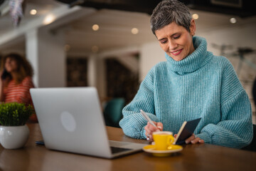 One senior woman in blue sweater sitting, working on laptop and writes in a notebook, modern business senior woman casual concept 