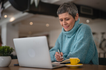 One senior woman in blue sweater sitting, working on laptop and writes in a notebook, modern business senior woman casual concept