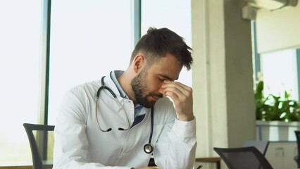 Stressed frustrated doctor wearing white uniform with stethoscope covering face with hands, sitting at work desk in hospital, sad young gp feeling desperate and guilty for medical failure