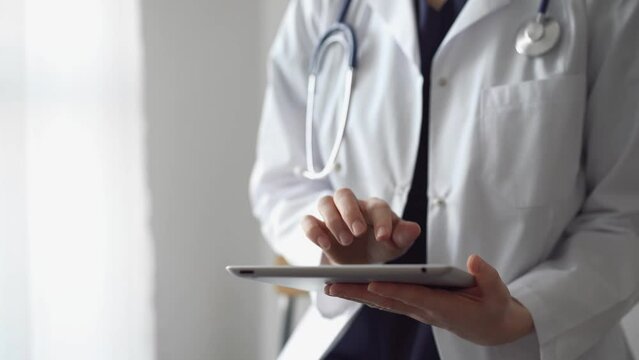 Doctor and patient sitting at the white desk near flair window in clinic. Unknown female physician use tablet computer for filling up medical record form. Medicine concept
