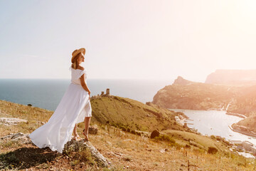 Happy woman in a white dress and hat stands on a rocky cliff above the sea, with the beautiful...