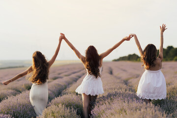 Young girlfriends in a white dresses standing with hands up in the middle of a lavender field in...