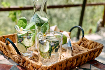 Refreshing summer drink - cucumber infused water with ice, sage, cucumber and lemon blossom on the table in the garden. Fresh healthy cold detox beverage. Fitness drink.