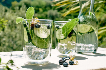 Refreshing summer drink - cucumber infused water with ice, sage, cucumber and lemon blossom on the table in the garden. Fresh healthy cold detox beverage. Fitness drink.