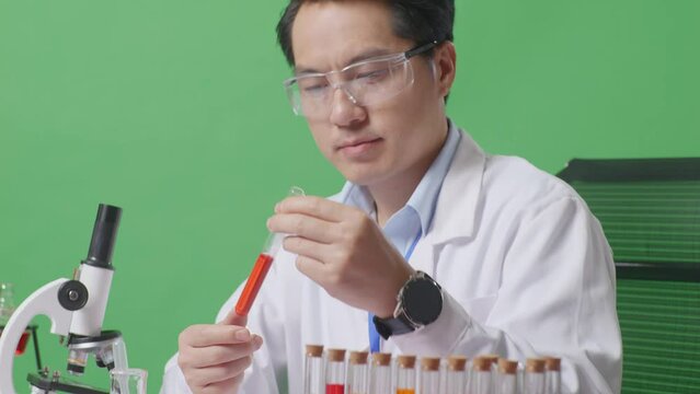Close Up Of Side View Of Asian Man Scientist Looking At Test Tube Then Thinking, Looking Around, And Raising Index Finger While Working On The Table With Microscope In The Green Screen Background Labo