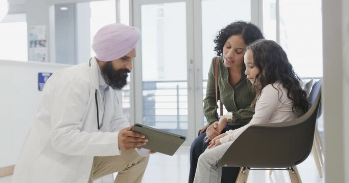 Biracial sikh doctor in turban with tablet talking to girl with mother in hospital, slow motion