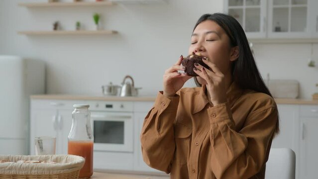 Moving camera shot Asian korean chinese hungry woman girl in kitchen eat fresh bakery cake sweet chocolate cupcake with nuts enjoy tasting brownie at home food delivery eating muffin end diet pleasure
