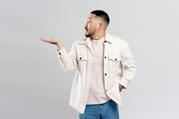 Surprised attractive asian man pointing with hand at copy space, isolated on gray background