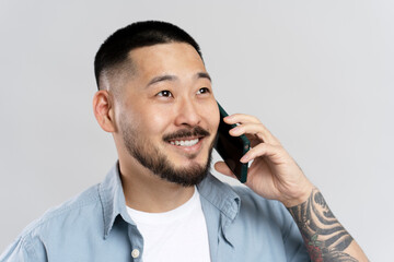 Portrait of handsome Asian man with stylish tattoo talking on mobile phone, isolated on gray background. Successful businessman answering call looking away 