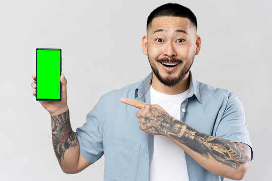 Excited Asian man holding mobile phone in hand, pointing finger at green screen. Attractive Korean male shopping online standing isolated on gray background
