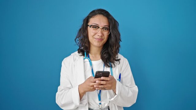 Young beautiful hispanic woman doctor smiling using smartphone over isolated blue wall background