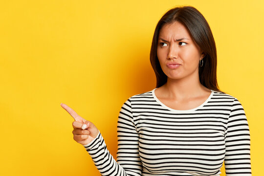 Beautiful woman stands on yellow background has long black hair wearing striped long sleeve t-shirt, frowing points finger to the side, copy space, time to think concept