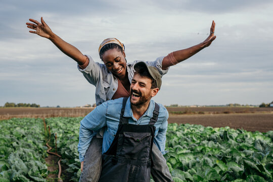 Smiling embraced diverse working couple having a piggyback ride and fun while spending time on their farm together.