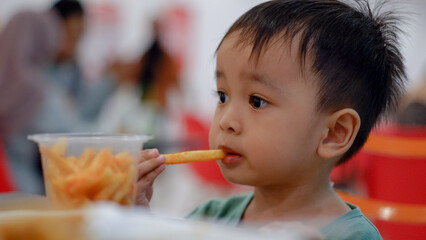 Cute Asian child eating French fries sitting at the dining table. Unhealthy kids foods concept.