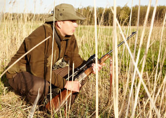 Fototapeta premium Hiding, game hunter and gun of man in nature on a safari for shooting and animal spotting. Sun, outdoor and sniper with scope and male person with a hunting weapon in Africa with patience and crouch
