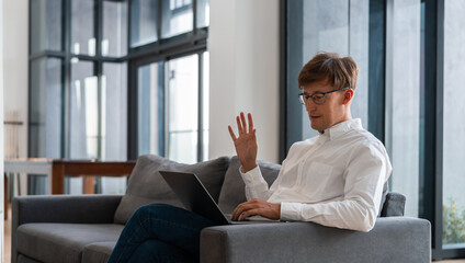 Smiling businessman sitting on couch and waving hand, video call