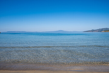 Fototapeta na wymiar Sandy empty beach transparent wavy sea water clear blue sky background. Greek seascape, sunny day.