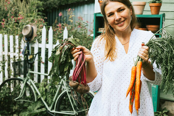cute farmer plus size real woman in casual white dress on veranda of country village house with ripe carrots and beets vegetables, eco-friendly food from garden, rich autumn harvesting