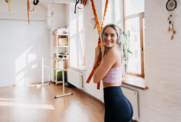 Gorgeous blonde caucasian young woman in sportswear stands at fitness club toothy smiles  looks at camera holds try straps at fitness club. Healthy lifestyle, workout. Happy American girl in earphones