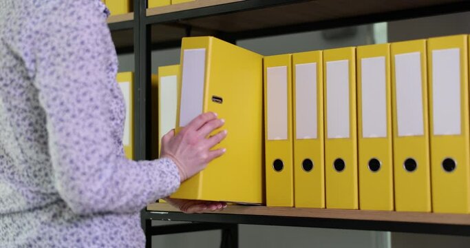 Woman employee putting back yellow ring binder with accounting papers to office archive. Organized folders structure with materials on shelf of rack