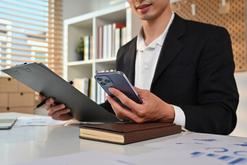 Select focus on hand, Young businessman holding smartphone and clipboard sitting at working desk