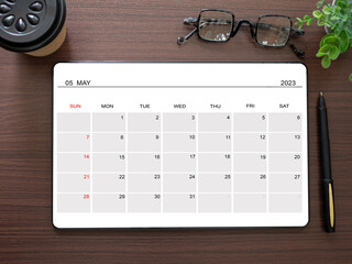 Top view, tablet showing May 2023 calendar app page and pen, tree, glasses, coffee cup on wooden...