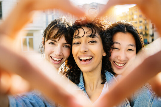 Three happy diverse young women drawing heart shape with hands outdoors. Smiling portrait of multi ethnic female friends having fun at city street. Love, youth lifestyle and friendship concept