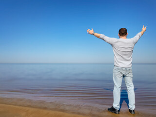 A man stands on the seashore with his arms outstretched. Rear view. Comfortable clothes. A person is resting in nature. A sunny, summer day. A happy, relaxed person. Copy space