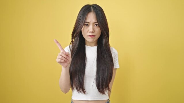 Young chinese woman saying no with finger over isolated yellow background