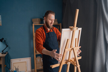 portrait caucasian bearded male artist in an apron in a home studio stands near an easel holds a...