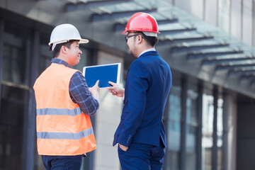 Two Industry Engineers Stand in modern building construction, Use Digital Tablet, Have Discussion. Engineers wearing safety vest and hard hat. 