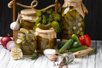 Pickled cucumbers in glass jars and spices and vegetables for preparation of pickles on wooden background.