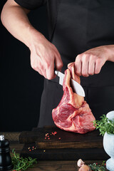 Chef cutting steak beef. Mans hands hold raw steak Tomahawk on rustic wooden cutting board on black...