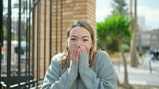 Young blonde woman standing with surprise expression covering mouth with hands at street