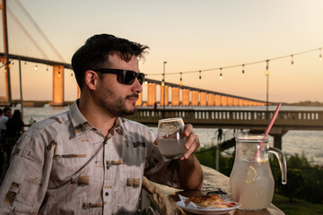 Hombre apuesto tomando limonada en un bar cerca del río, al atardecer