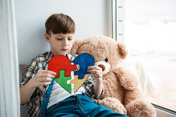 Lonely little boy with teddy bear sitting at home. A little Autistic child holds a heart with puzzle pieces in his hands. World Autism day