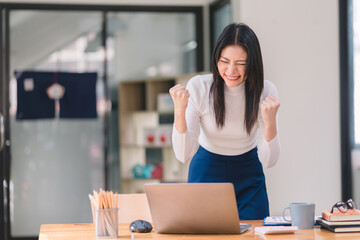 Happy and excited, a young asian woman student or office worker, celebrates achieving her goal of winning an online competition or receiving good news via email by raising hands in euphoria.