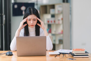 The stressed and exhausted millennial Asian businesswoman is seen sitting at her office desk with her hand on her head, indicating a hard working day where she is overloaded with work. - obrazy, fototapety, plakaty