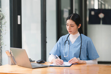 A concentrated female doctor or nurse is working online with a laptop while sitting at a desk in a hospital consultation room. medicine, technology and healthcare concept