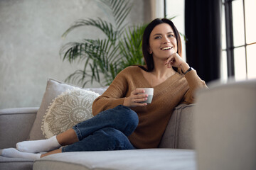 Happy dreamy middle aged woman sitting on comfortable sofa in living room with cup of black tea or coffee
