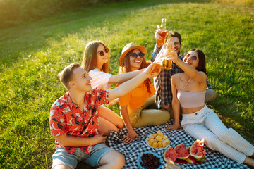 Cheerful company on sunny meadow at picnic is resting, drinking beer, cheers. Stylish friends enjoy sunny day on beautiful green meadow. Vacation, picnic, friendship or holliday concept.