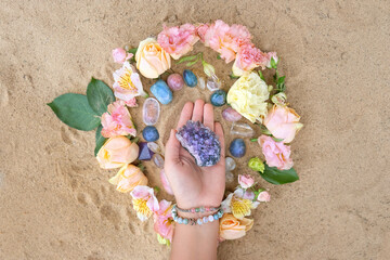 Hand with amethyst druse, gemstones and flowers on sand natural background. Crystal Ritual,...