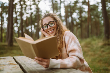 Obraz premium Happy young woman is sitting at table with book in beautiful forest. Beautiful woman is enjoying fresh air in forest reading book. Concept of relaxation, solitude with nature, lifestyle.