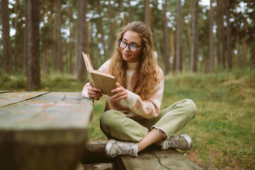 Happy young woman is sitting at table with book in beautiful forest. Beautiful woman is enjoying...