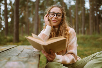 Happy young woman is sitting at table with book in beautiful forest. Beautiful woman is enjoying...