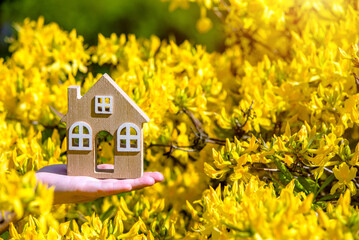 The girl holds the house symbol against the background of blossoming yellow rhododendron
