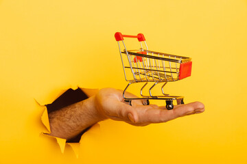 Male hand holds through a hole a mini grocery shopping trolley on a yellow paper background